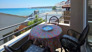A balcony with a table and chairs, overlooking a beach with boats and buildings in the distance. - ARİFE REZİDANCE NO.2 (Didim)