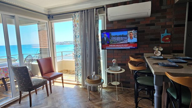 A spacious room with a large TV, wooden flooring, a ceiling fan, and a view of the sea through large windows. - ARİFE REZİDANCE NO.2 (Didim)