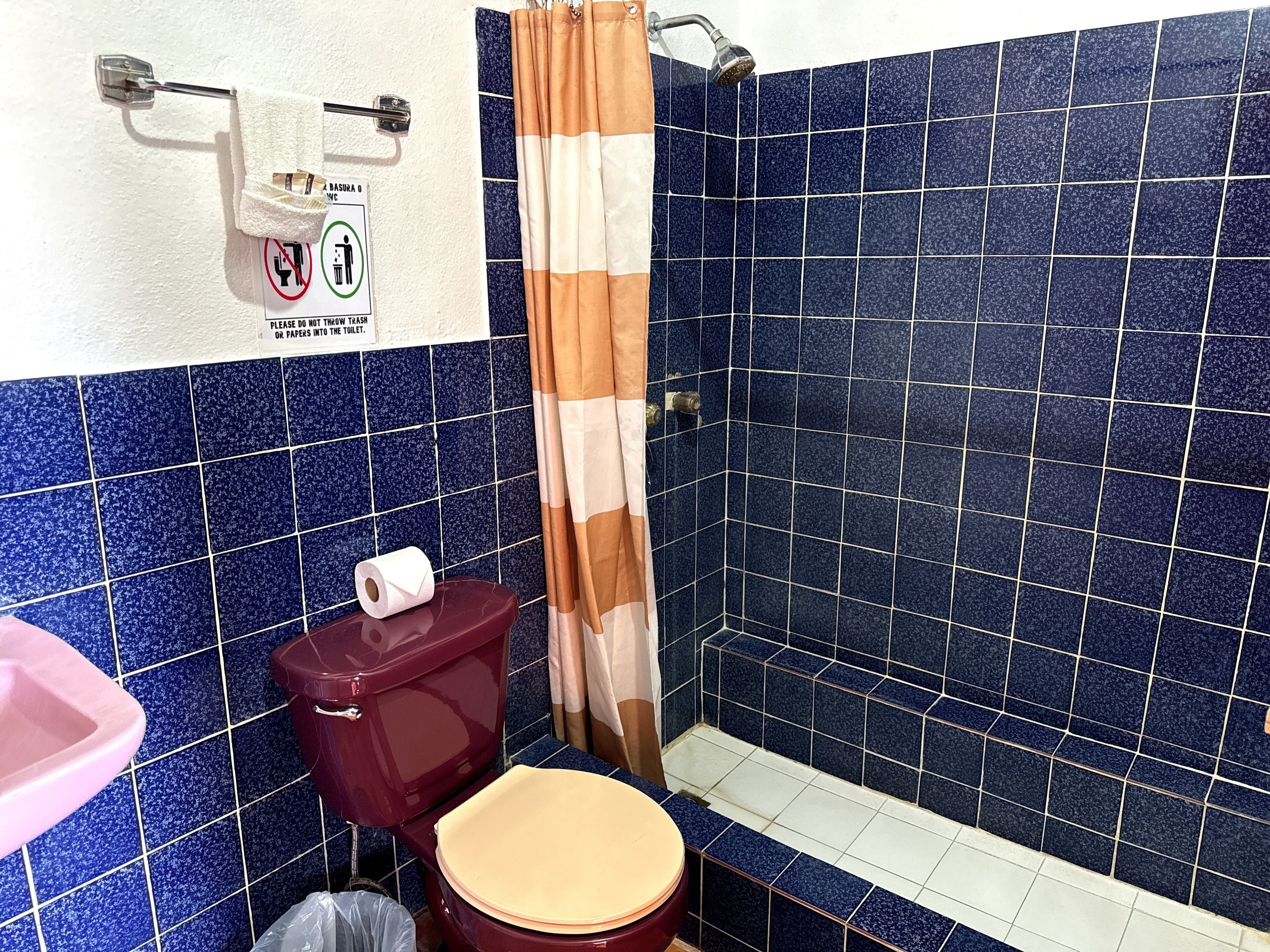 Egy fürdőszoba, melynek a falai kék csempével vannak burkolva, van benne egy piros WC, egy zuhanyfüggöny és egy kézfertőtlenítő-adagoló.