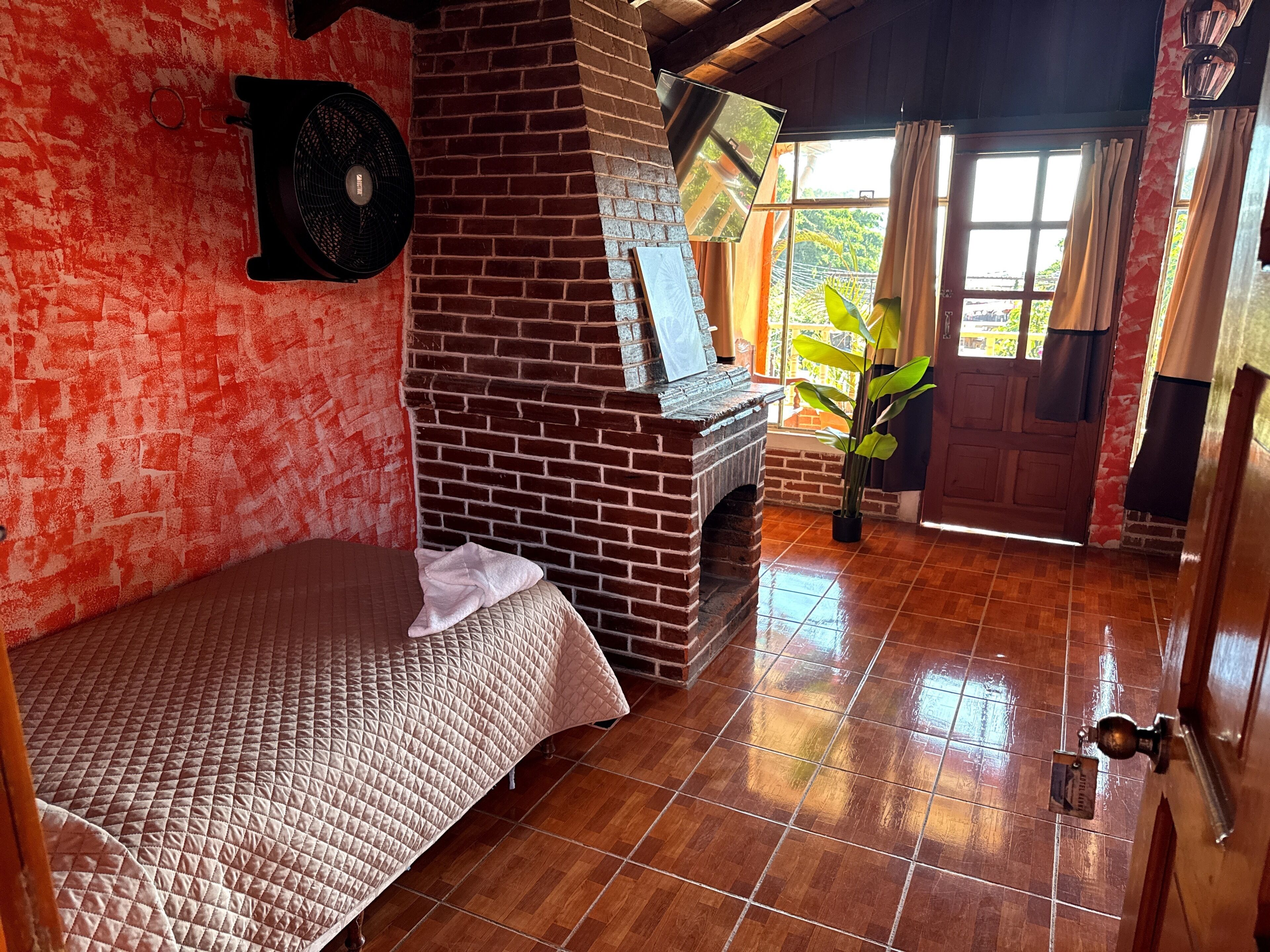 A room with a brick fireplace, a bed with a quilt, a fan, and a view of greenery outside.