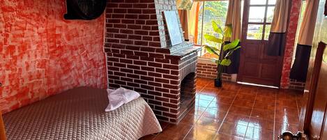 A room with a brick fireplace, a bed with a quilt, a fan, and a view of greenery outside.