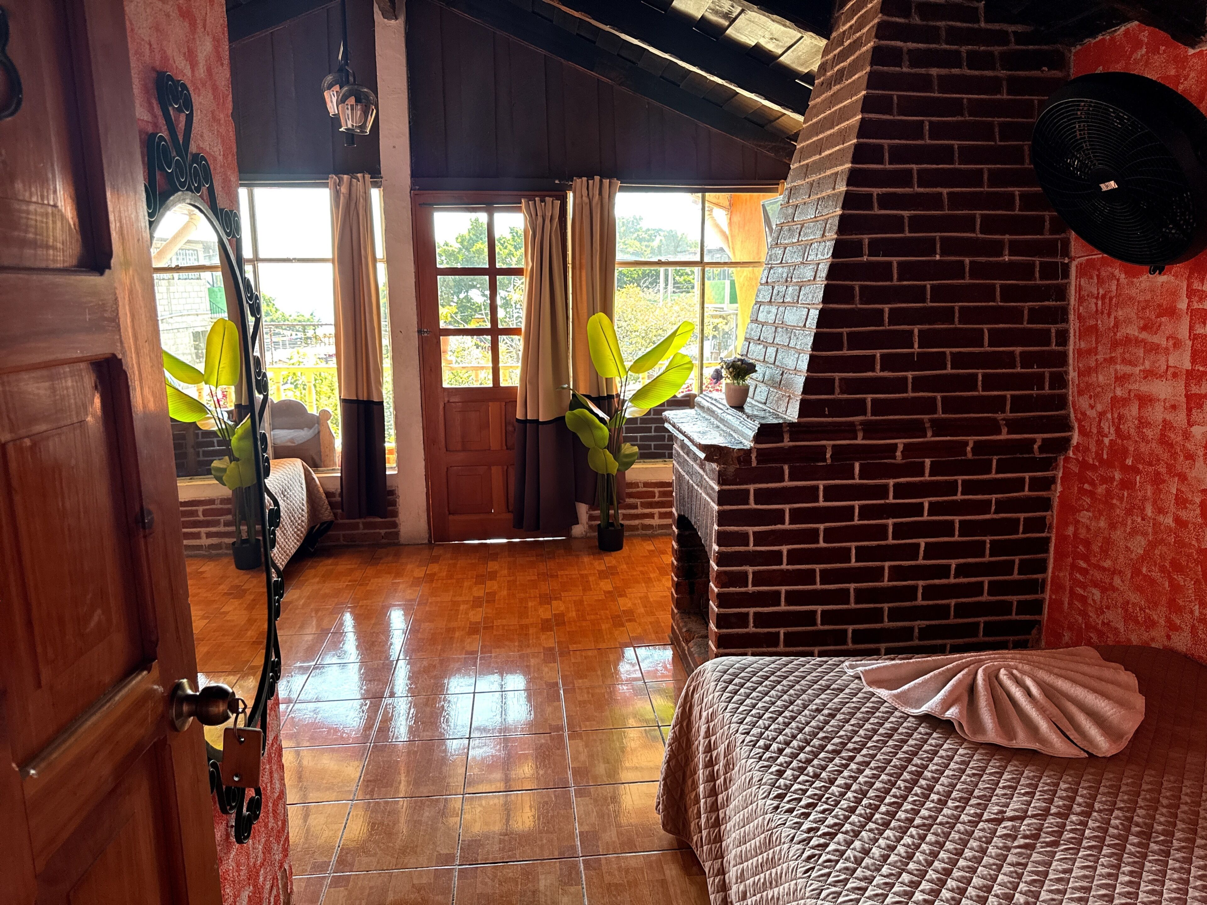 A room with a wooden door, a bed with a quilted cover, a brick wall, a window with curtains, and a potted plant.