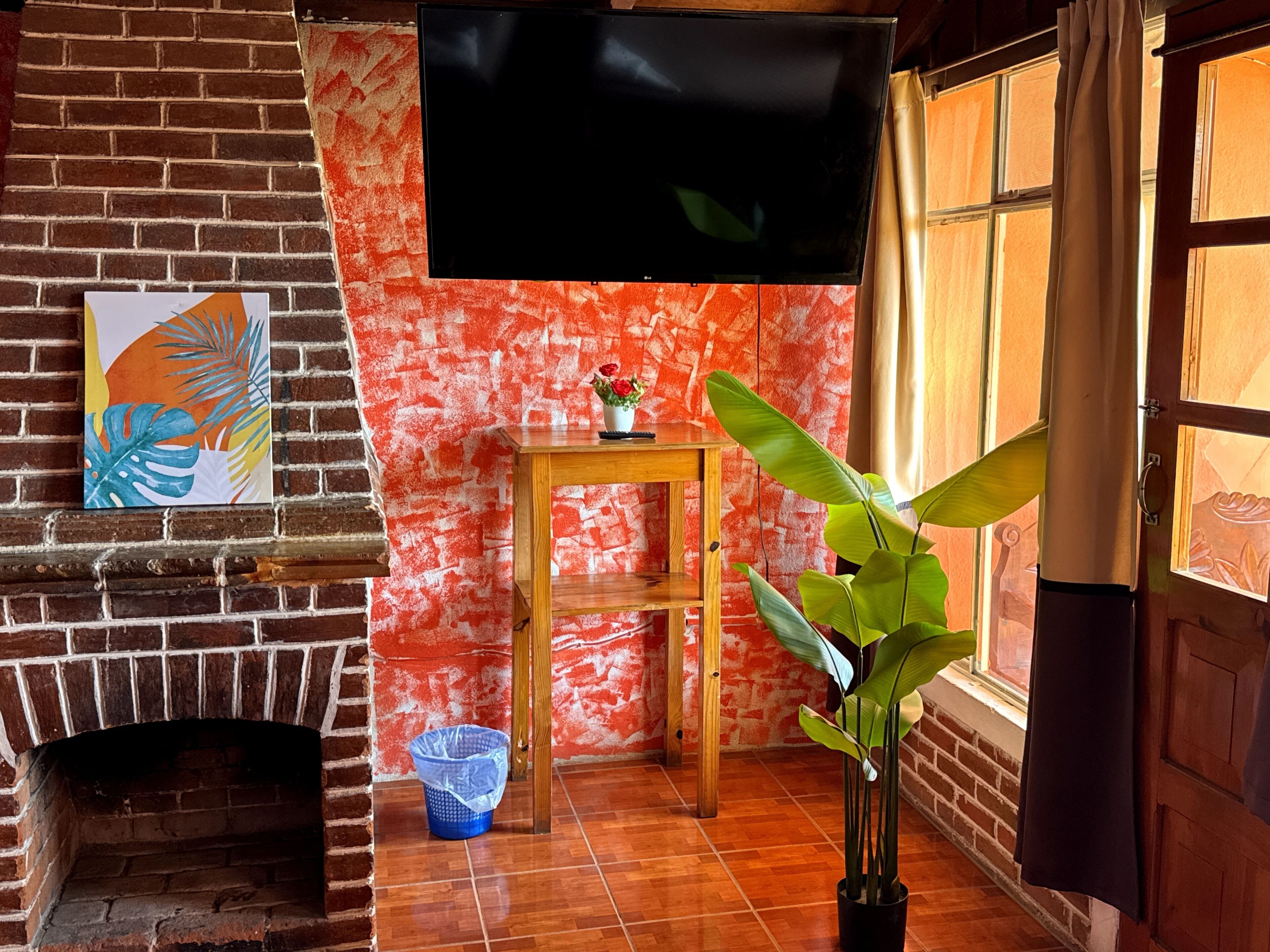 Een kamer met een haard van baksteen, een flatscreen televisie aan de muur, een houten tafel met een bloevas en een grote kamerplant.