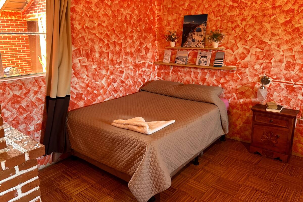 Een kamer met een bakstenen muur, een houten vloer, een bed met quilt, een nachtkastje, een plank met boeken en een foto, en een raam met gordijnen.