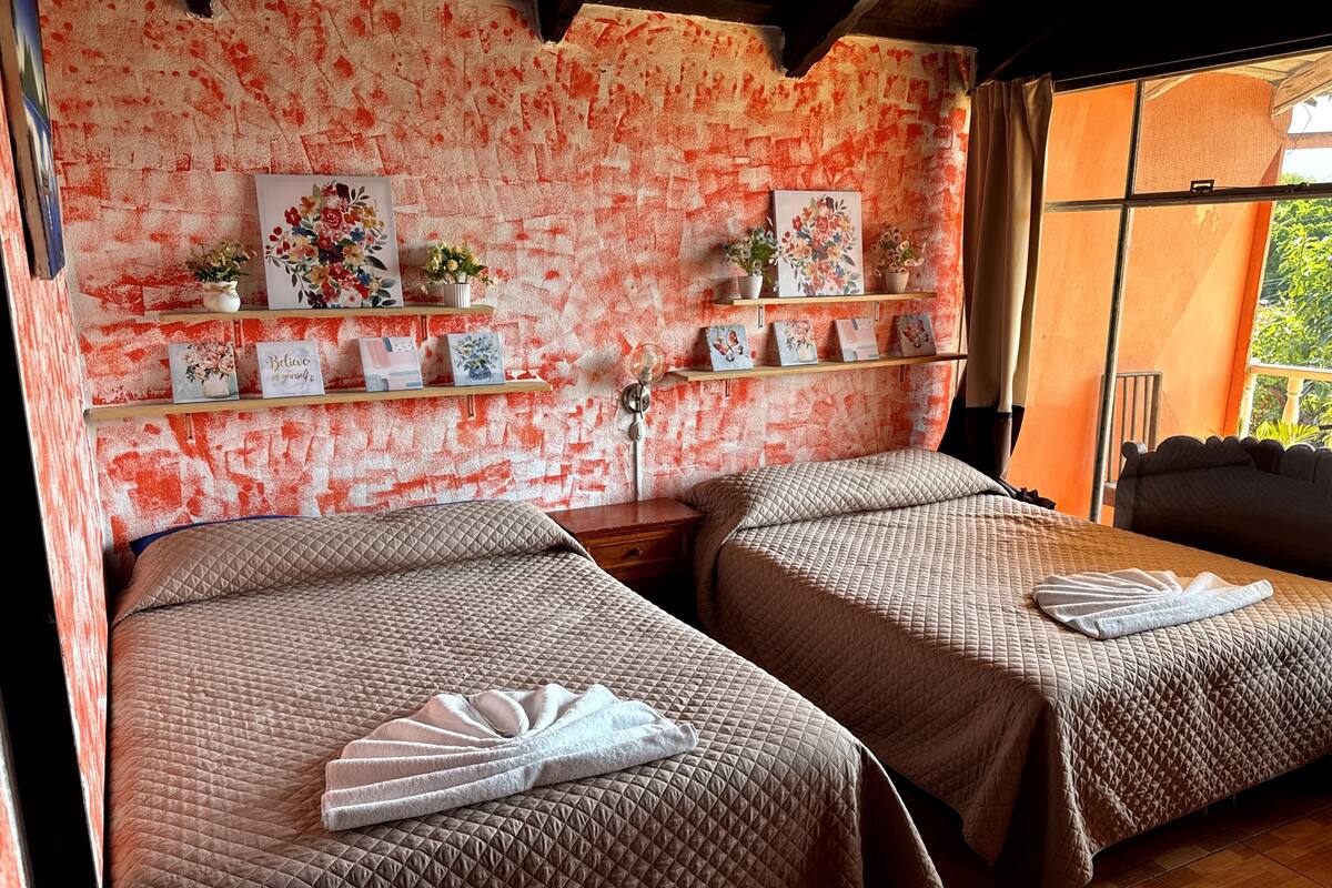 Twee bedden met gewatteerde dekbedden, een muur met een rood-wit patroon, en planken met ingelijste foto's en kleine potplanten.