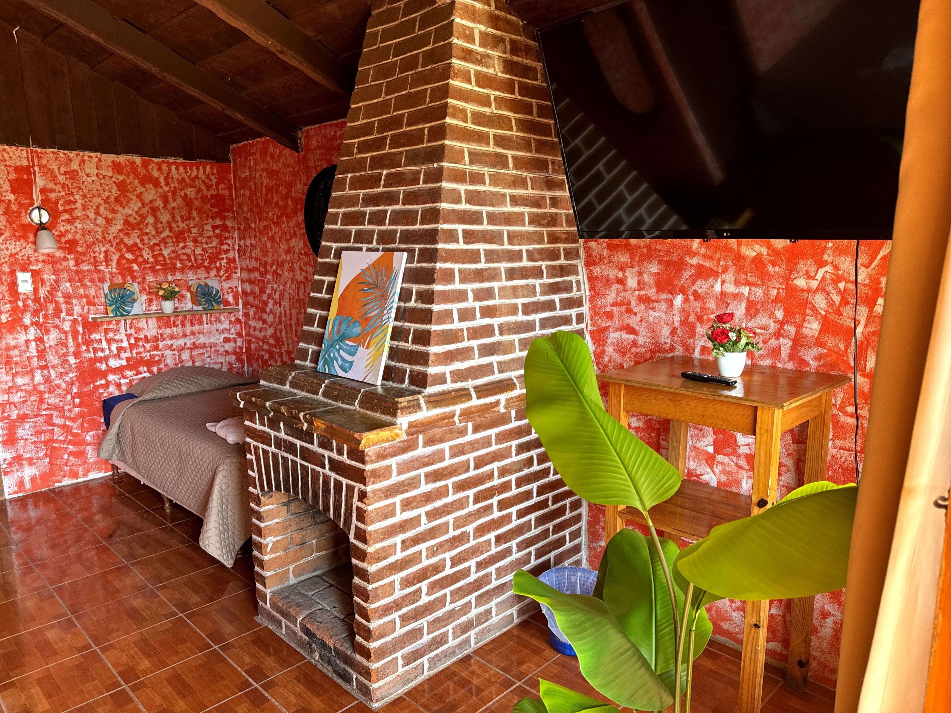 A room with a brick fireplace, a bed, a table with a plant, and a wall with a red textured pattern.