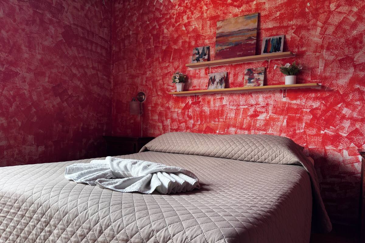Een slaapkamer met een bed, een rode, gestructureerde muur en een plank met kunstwerken en planten.