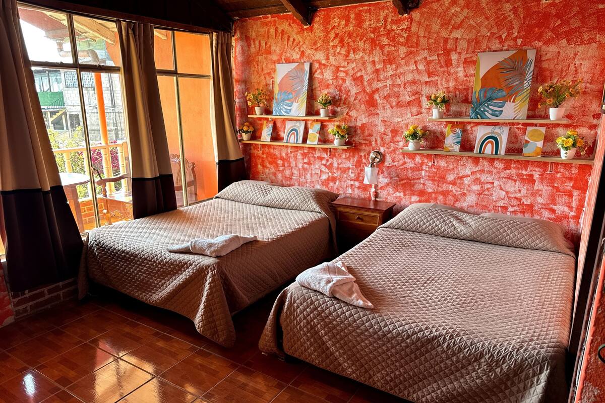 Een kamer met twee bedden, een raam met gordijnen, een rode, gestructureerde wand en een plankje met decoratieve spullen.