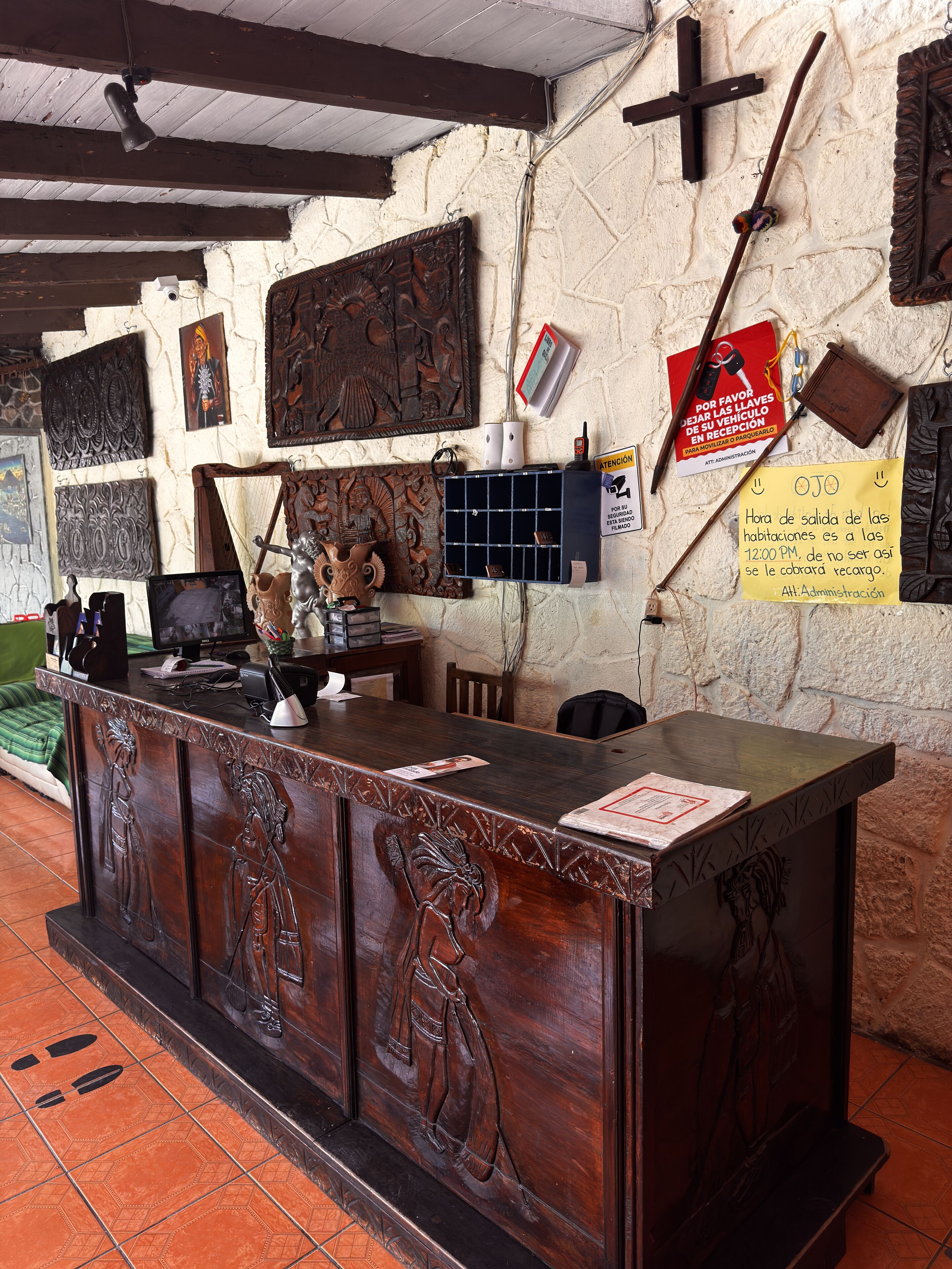 Bancada de recepção com painéis de madeira entalhada, uma cruz na parede e diversos elementos decorativos, incluindo uma moldura com fotografia e um aviso.