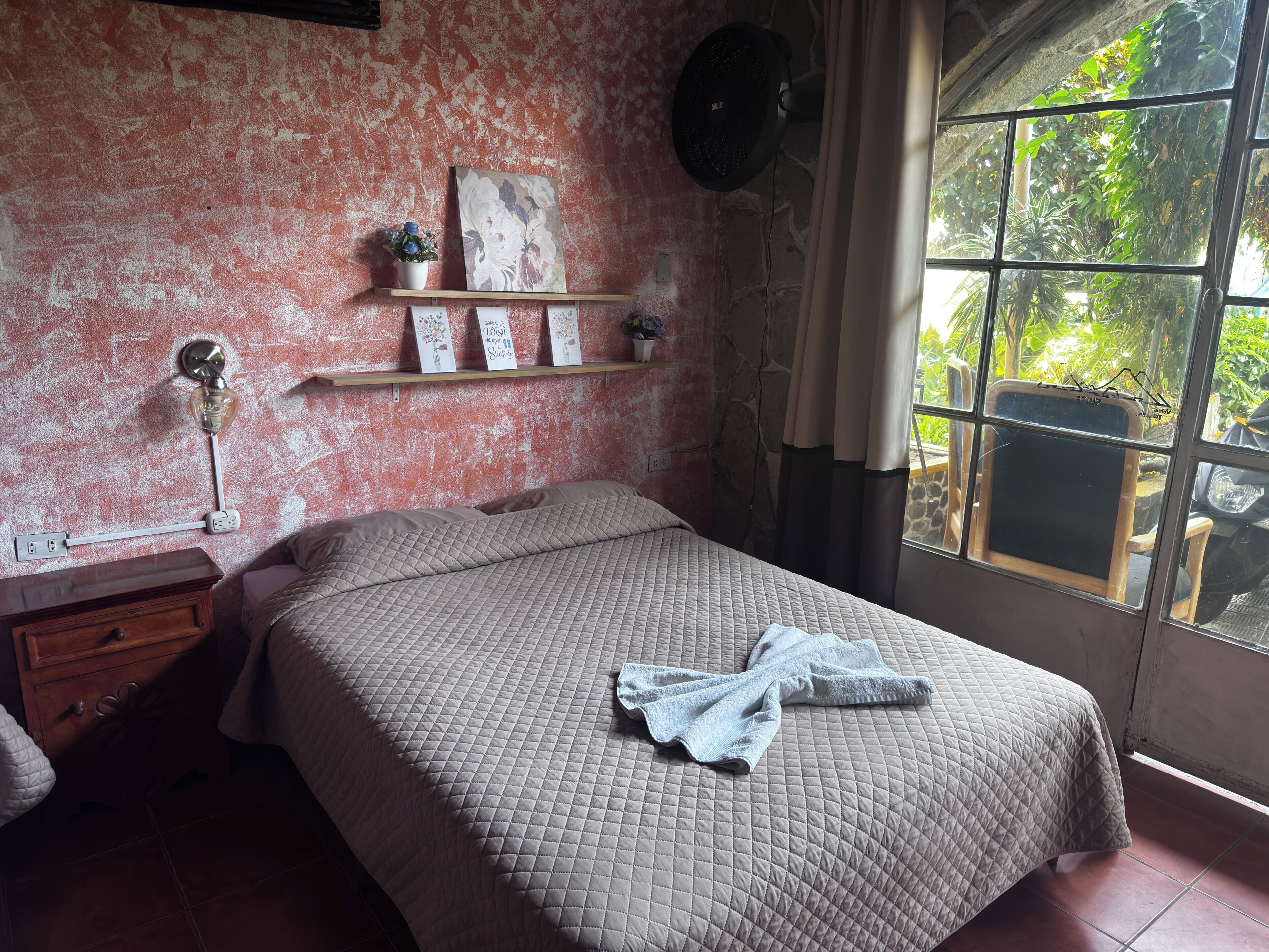 A bedroom with a bed, a nightstand, a wall-mounted lamp, a shelf with decorative items, and a window with a view of greenery.