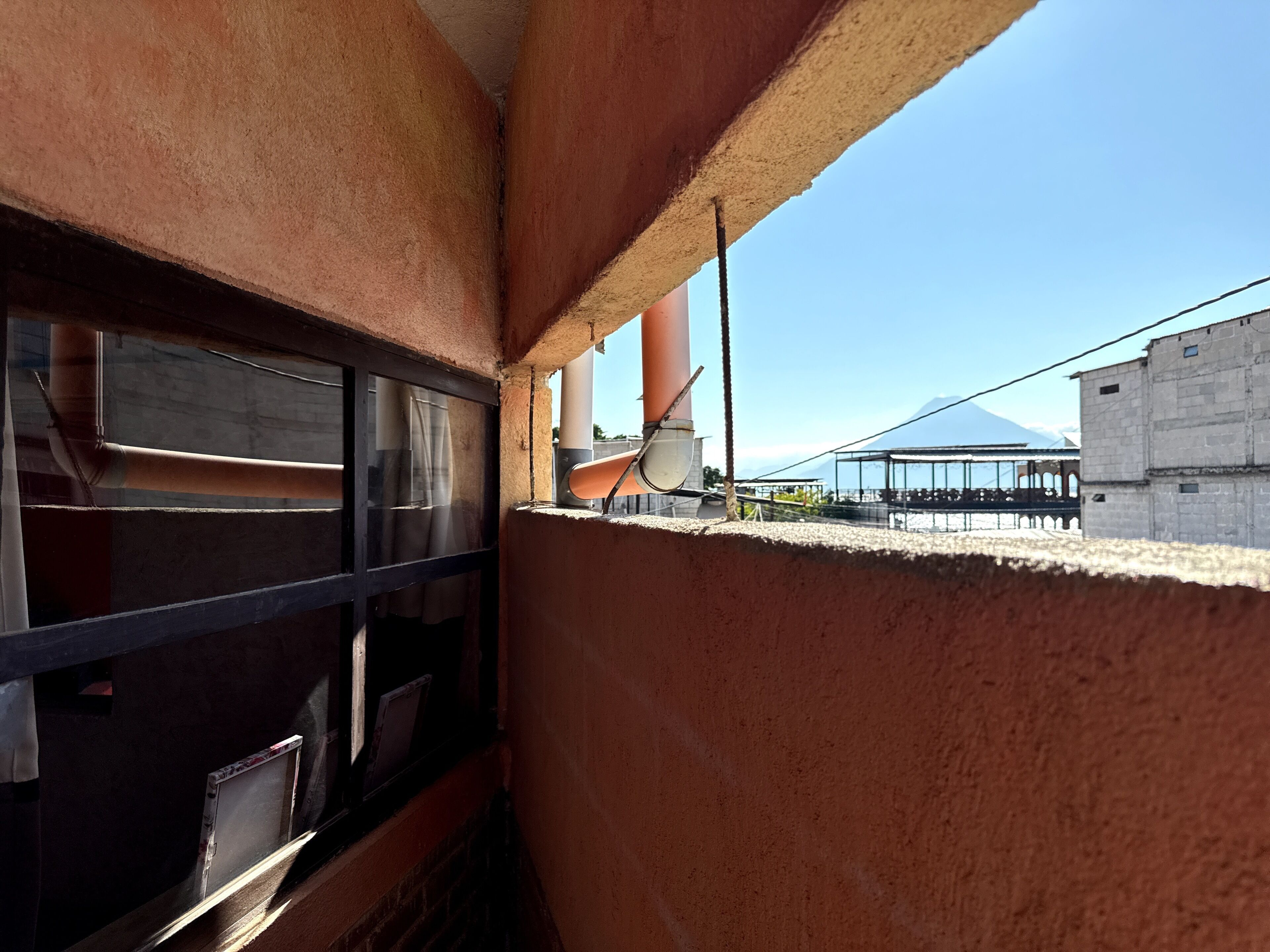 Un couloir avec un mur en béton, une fenêtre avec des stores et une vue sur un immeuble sous un ciel dégagé.