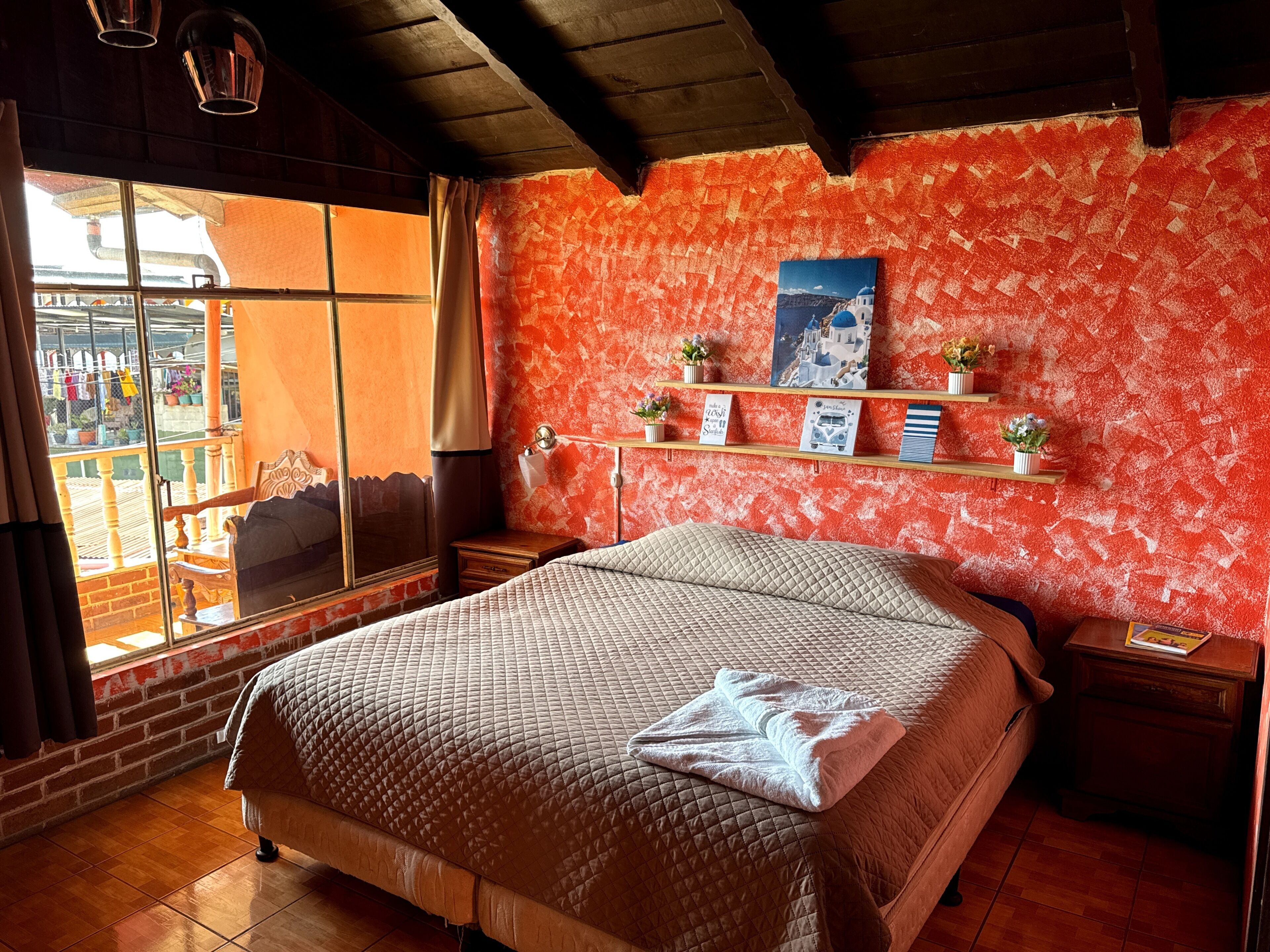 A bedroom with a large bed, a window with a view of the outside, a shelf with decorative items, and a ceiling fan.