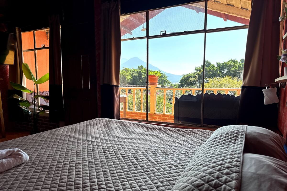 Een kamer met een bed, een raam met uitzicht op groen en bergen, en een balkon met een hekwerk.