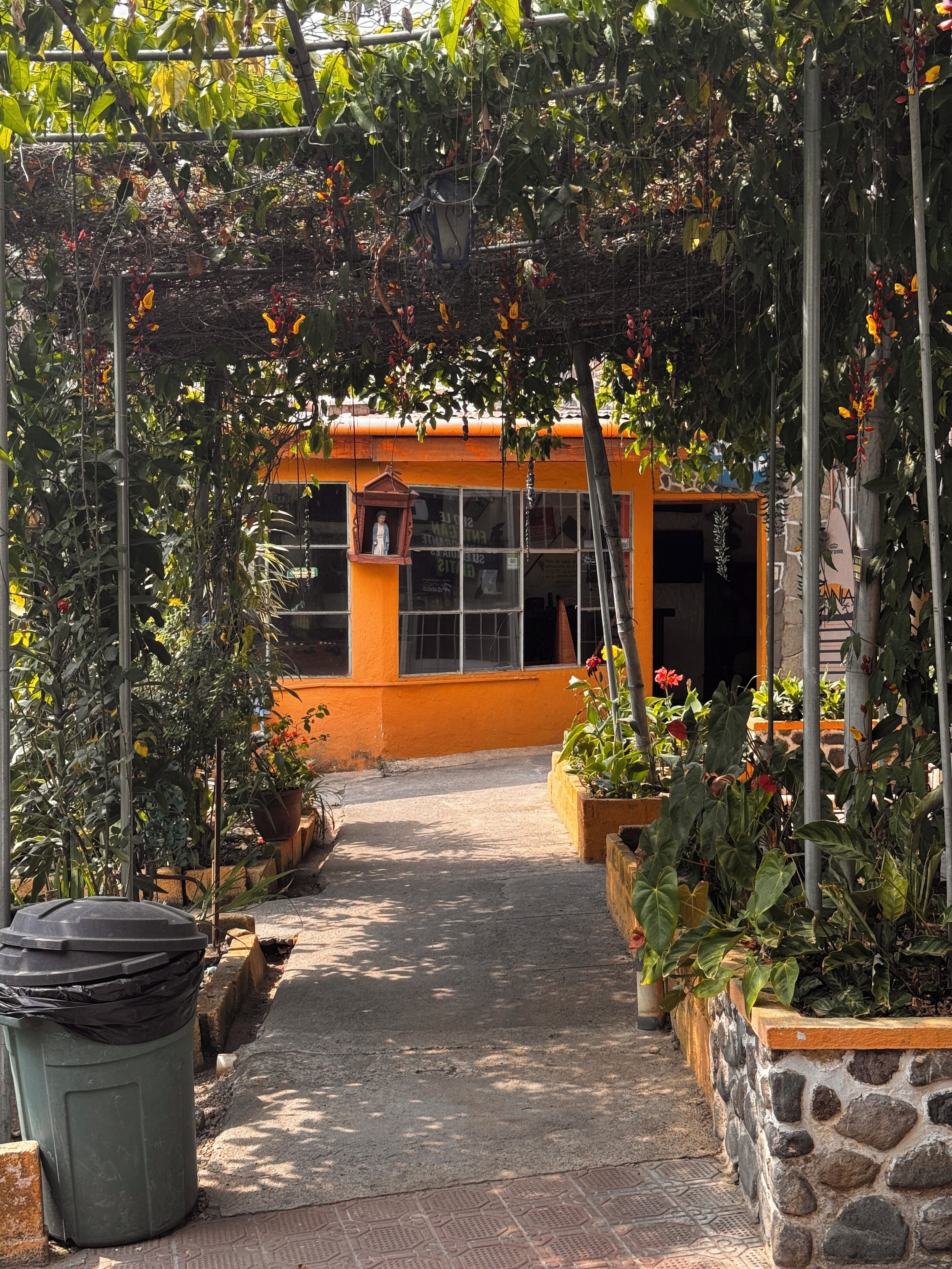 Un camino conduce a un edificio de fachada amarilla, rodeado de plantas en macetas y un farol colgante.