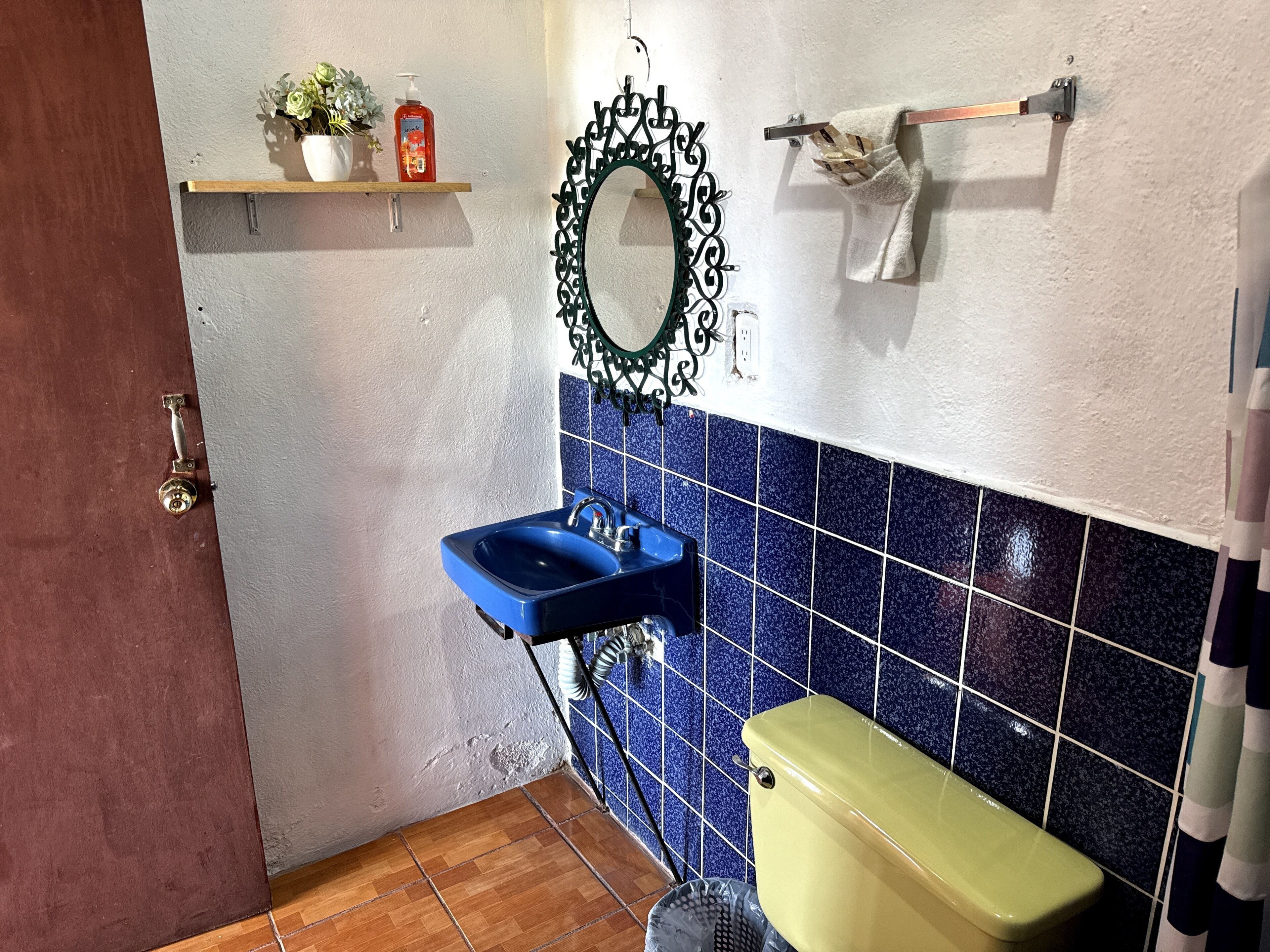 Casa de banho com lavatório azul, sanita amarela, espelho decorativo e uma estante com uma planta em vaso e uma garrafa.