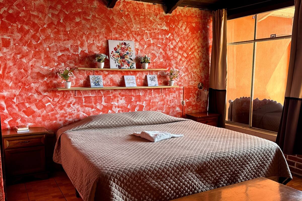 Een slaapkamer met een gewatteerde spreideken, een houten nachtkastje, een bijzettafeltje, een raam met gordijnen en een muur met een rood reliëfpatroon.