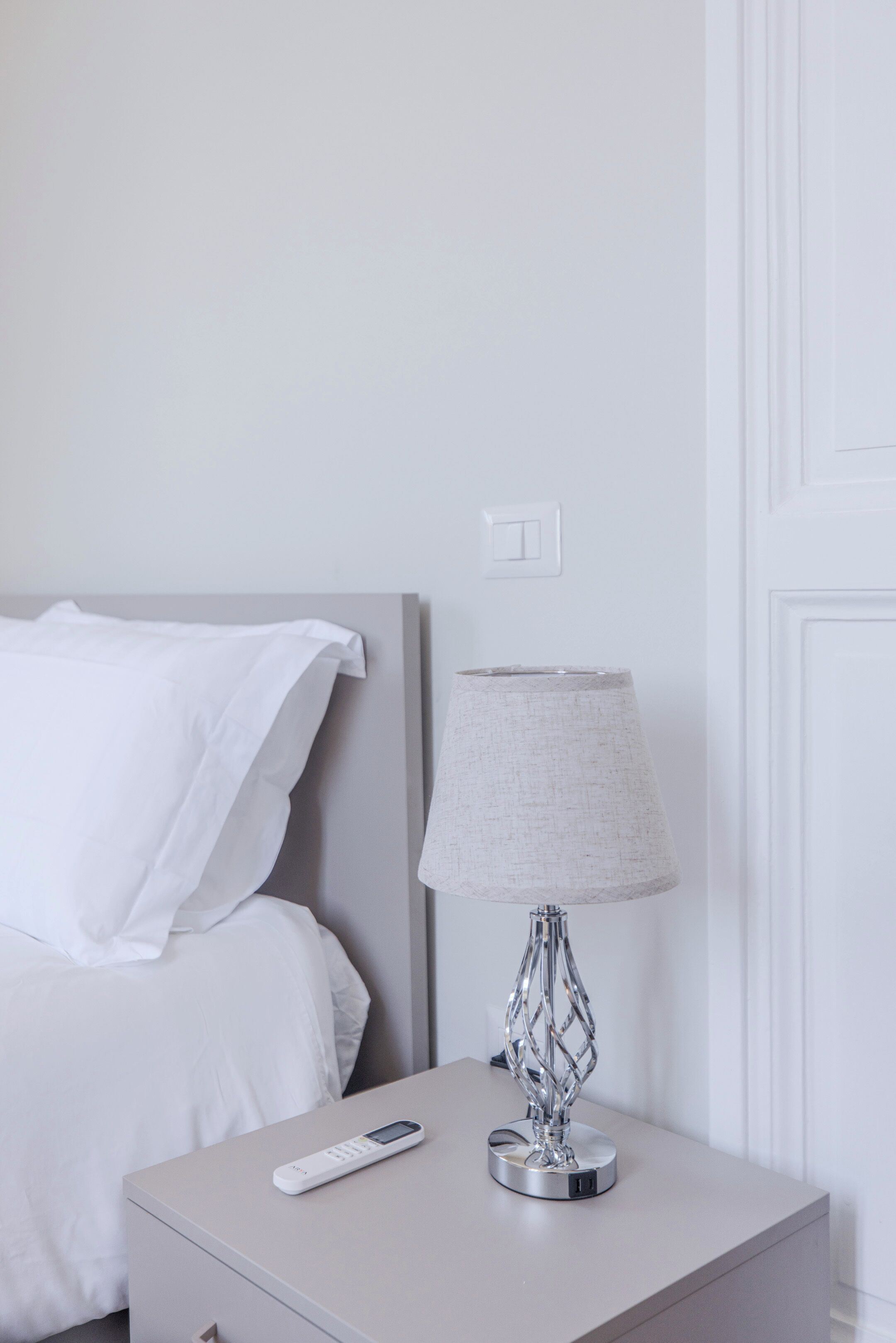 A neatly made bed with white linens, a bedside table with a lamp, and a remote control on the table.