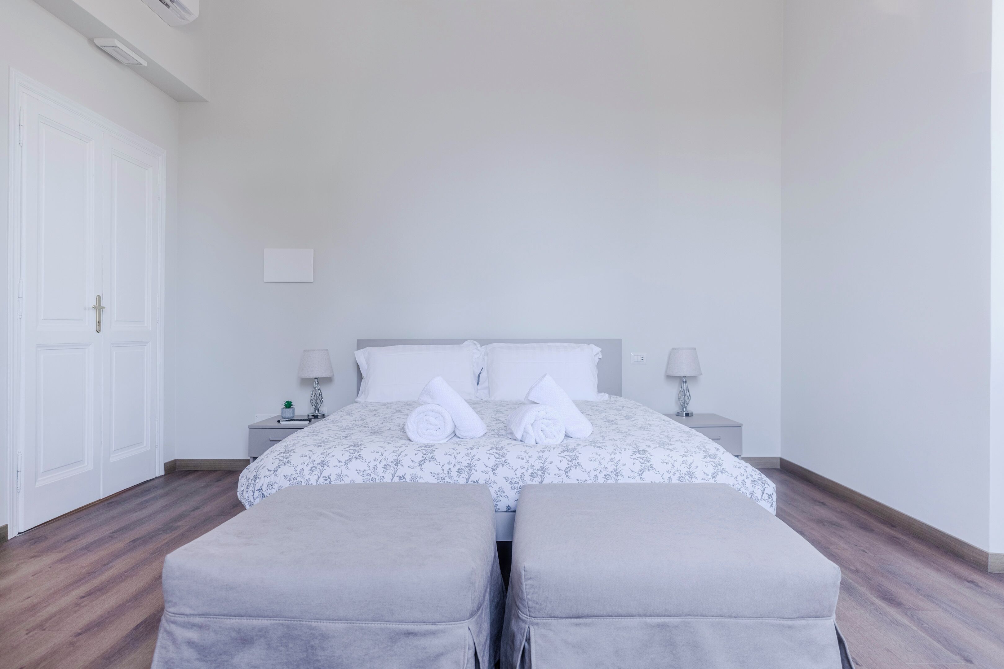 A hotel room with a bed, two bedside tables, a nightstand, a door, and two grey ottomans.