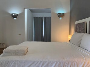A hotel room with a bed, a nightstand, a lamp, and a view of a corridor with a door. - Tracadero Marina 40 Apt, 1 Bdr. Sea View (Dominicus)