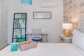 A hotel room with a bed, a desk with a chair, a nightstand with a lamp, and a wall-mounted air conditioner.