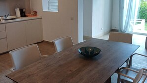 Et moderne køkken med hvide skabe, et spiseområde med et træbord og hvide stole samt udsigt til en have gennem et stort vindue.
