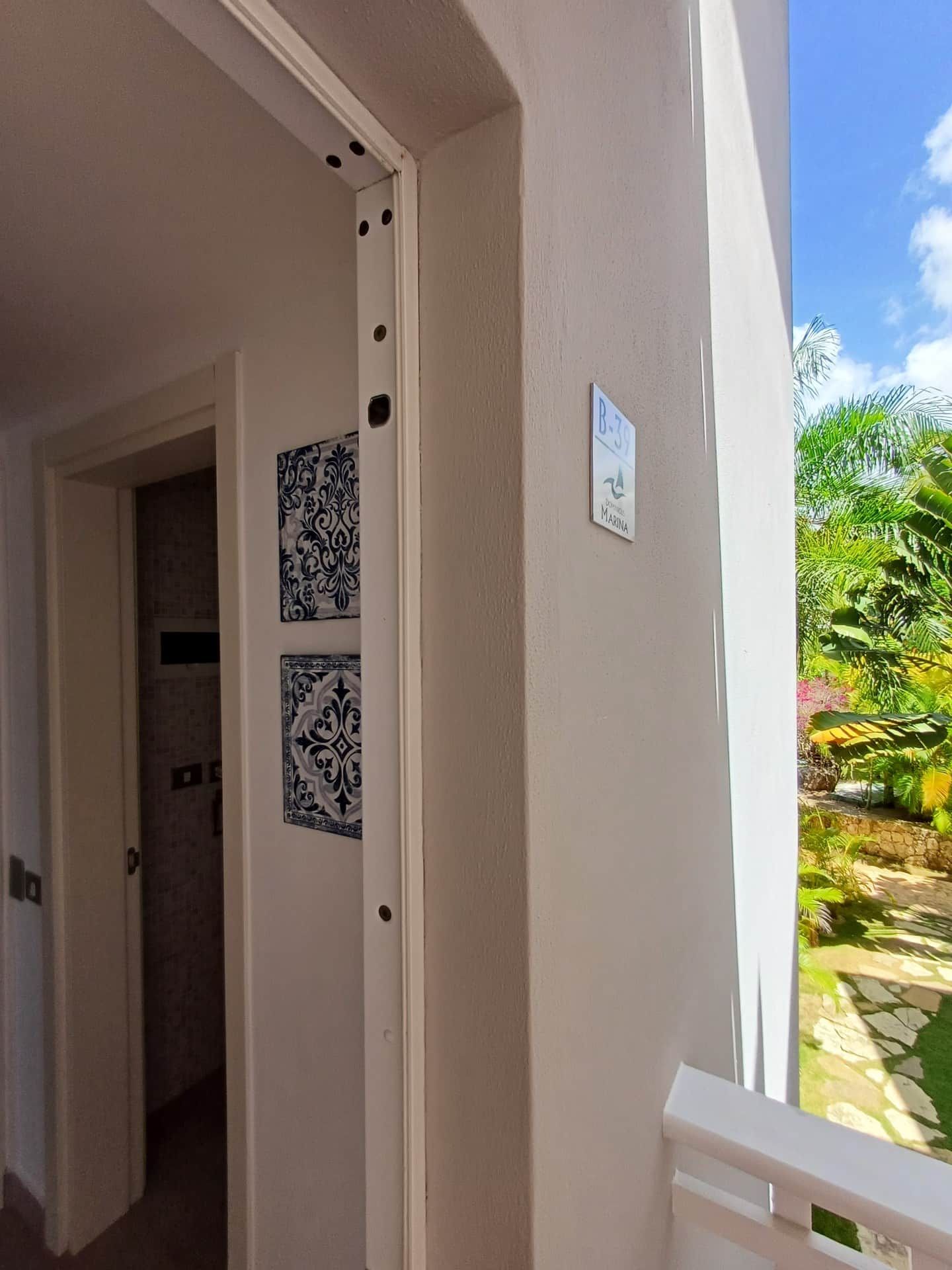 A hallway with a door leading to a bathroom, a small sign with the number 'B-39', and a view of a tropical garden outside.