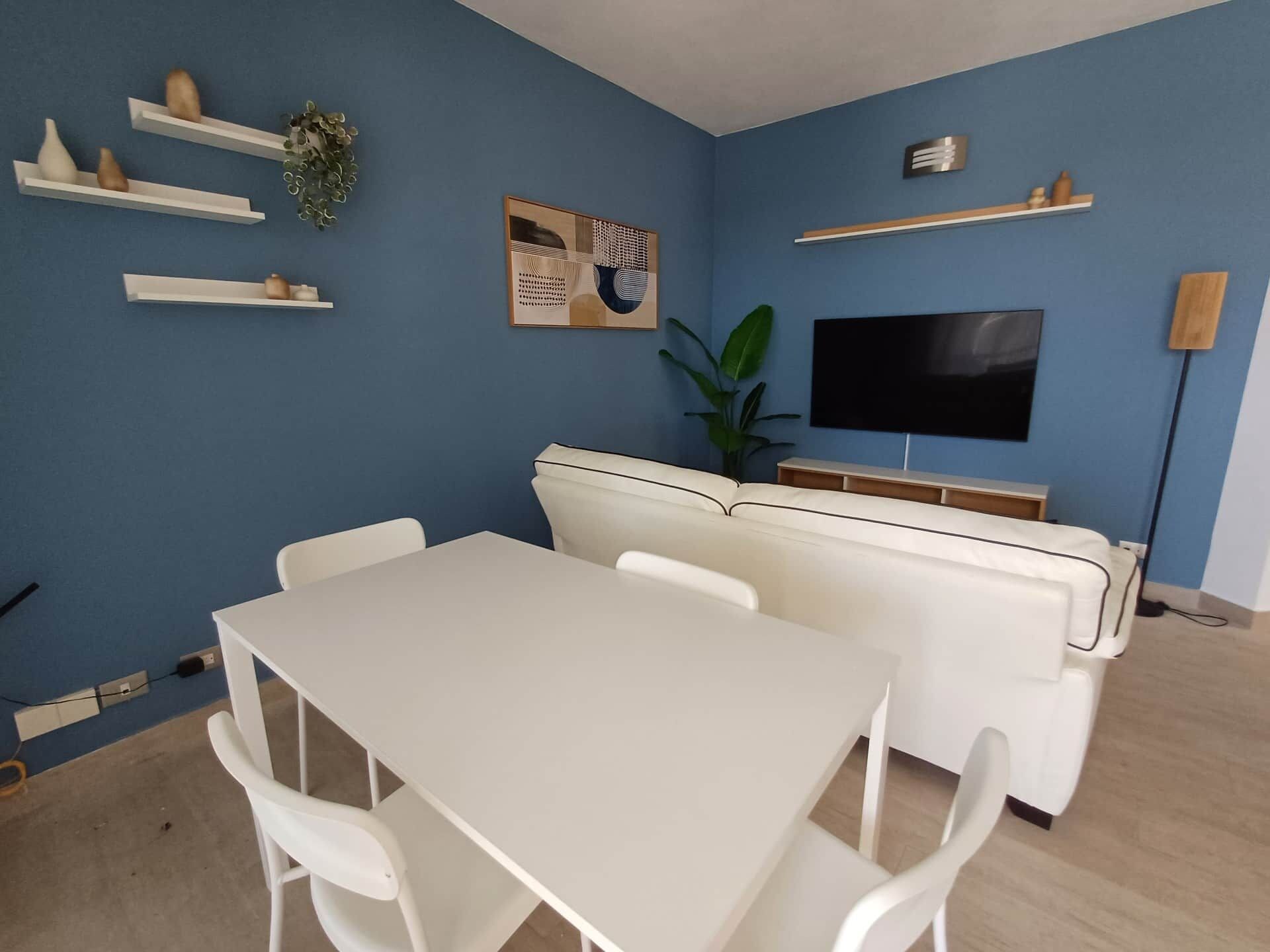 A living room with a white sofa, a TV mounted on the wall, and a dining table with chairs.