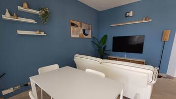 A living room with a white sofa, a TV mounted on the wall, and a dining table with chairs.