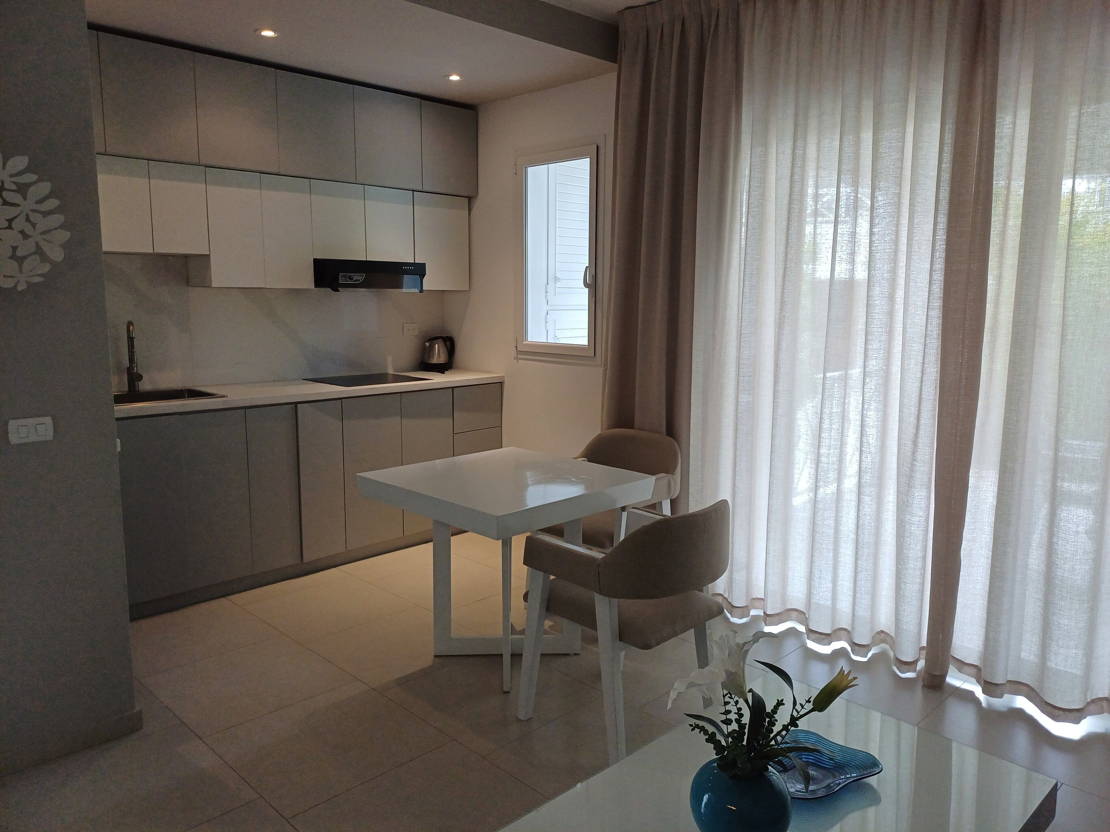 Una cocina moderna con una mesa y sillas blancas, una ventana con cortinas y un jarrón con flores sobre la mesa.