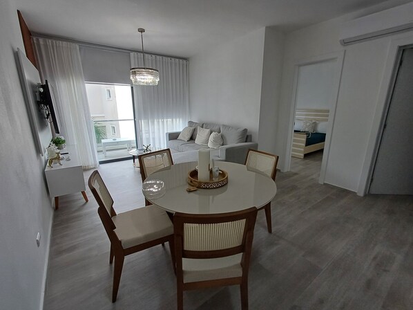 A modern living room with a dining area, featuring a round table, wooden chairs, a sofa, and a large window with curtains.