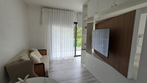 A modern living room with a sofa, a TV mounted on a wooden panel, and a view of greenery outside.
