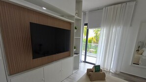 A modern living room with a large flat-screen TV mounted on a wooden panel wall, a sliding glass door leading to a balcony, and a seating area with a small table and cushions.