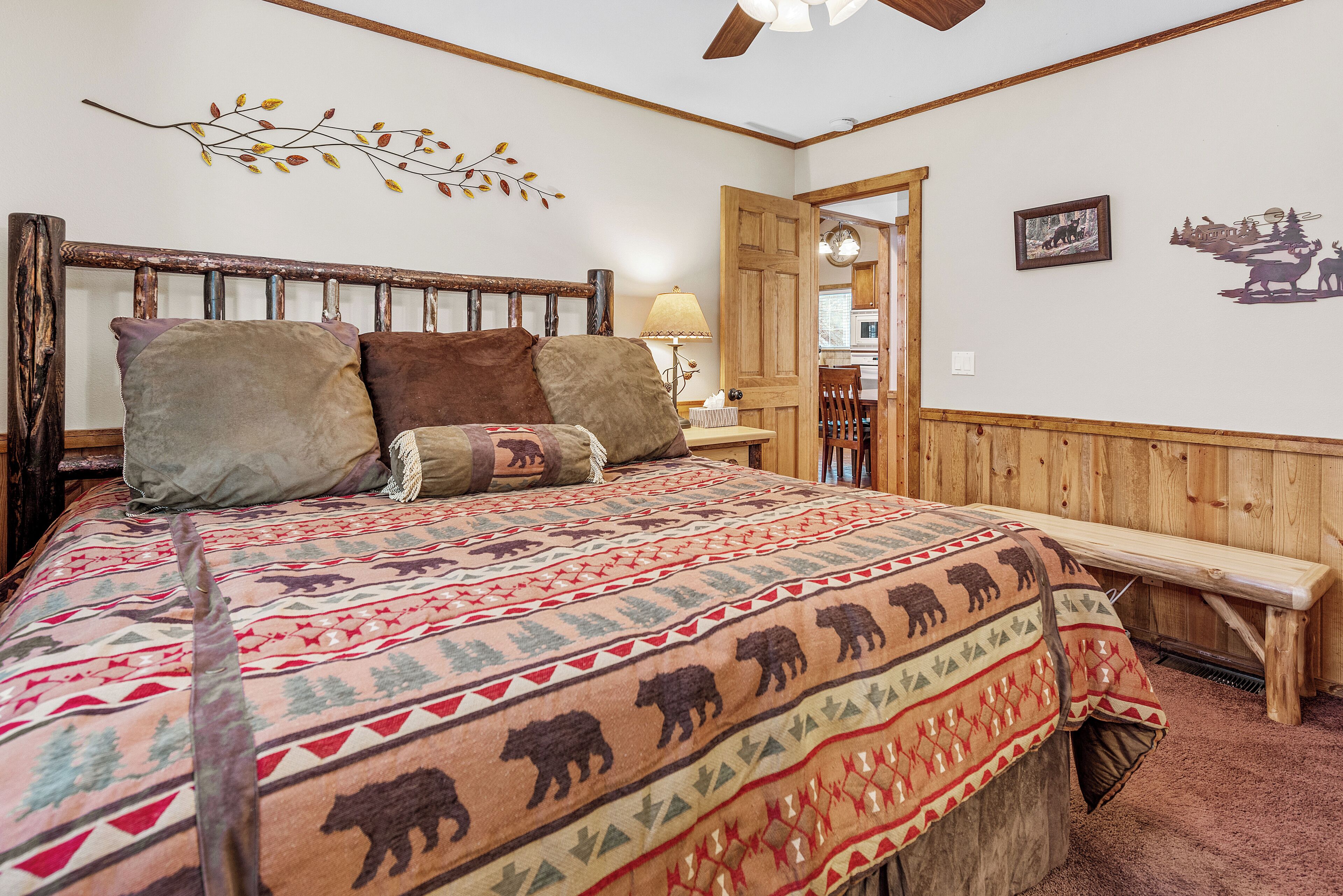 Un dormitorio con cabecera de madera, cama con manta estampada, un banco, una lámpara y una puerta que da a otra habitación.