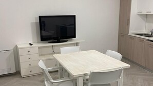 Une pièce moderne et minimaliste, avec une table et des chaises blanches, un téléviseur à écran plat posé sur un meuble blanc, et un coin cuisine doté de placards en bois.