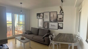 Uma sala de estar moderna com um sofá, uma mesa de jantar com cadeiras e uma mesa de centro. Há quadros nas paredes e uma vista pela porta de vidro.