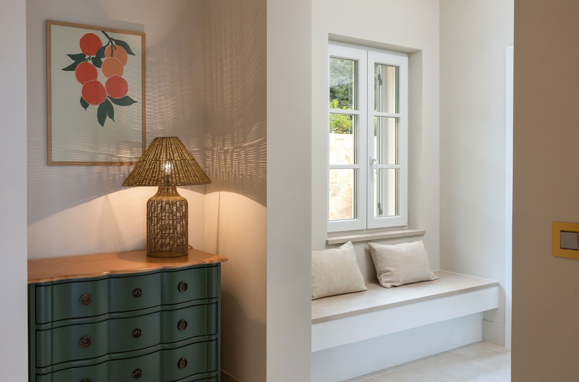A room with a green dresser, a wall-mounted lamp, a window with a white bench and cushions, and a framed artwork of fruit.