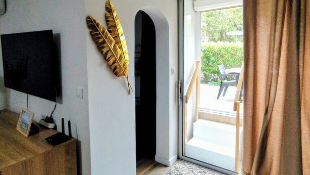 A modern living room with a flat-screen TV, a wooden cabinet, a decorative golden leaf, a glass door leading outside, and a brown curtain. - Magnificent Studio close to the sea. (Calvi)