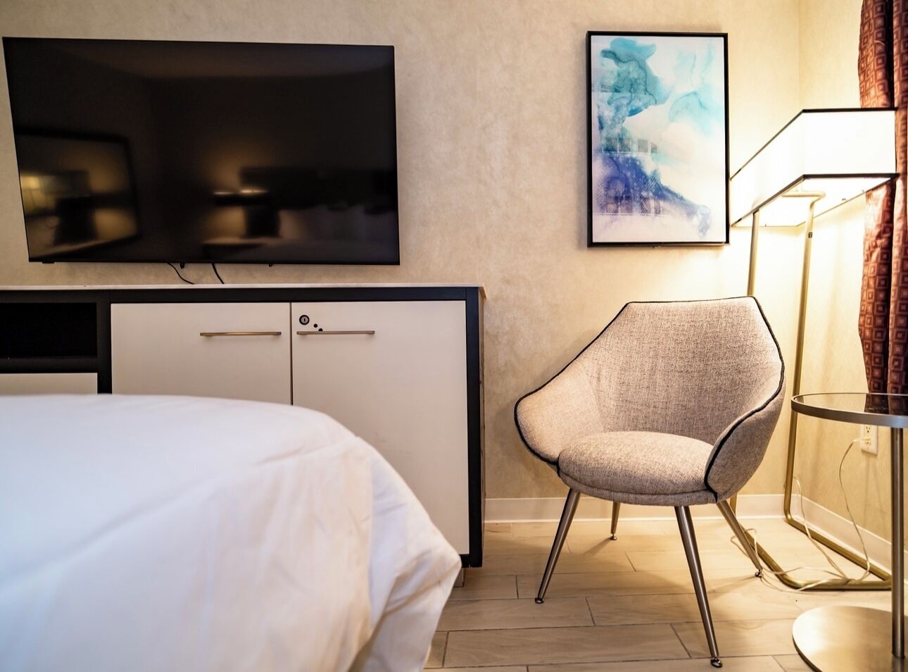 A hotel room with a bed, a desk with a flat-screen TV, a chair, and a lamp.