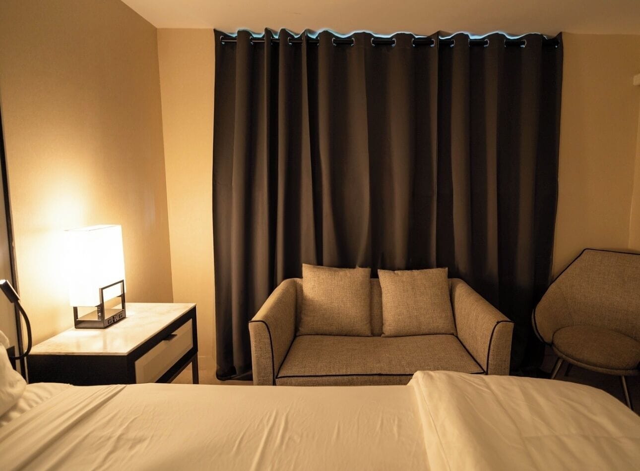 A hotel room with a bed, a sofa, a chair, a nightstand with a lamp, and a curtain.