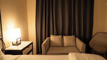 A hotel room with a bed, a sofa, a chair, a nightstand with a lamp, and a curtain.
