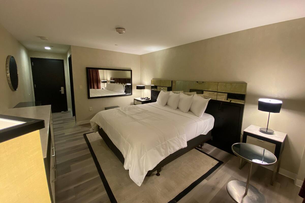 A hotel room with a large bed, a bedside table with a lamp, a mirror, and a door leading to another room.