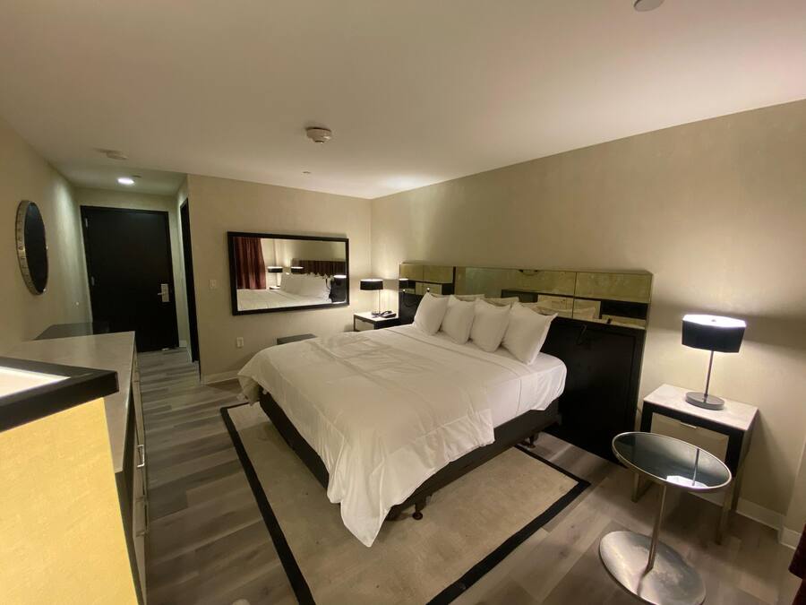 Een hotelkamer met een groot bed, een nachtkastje met een lamp, een spiegel en een deur naar een andere kamer.