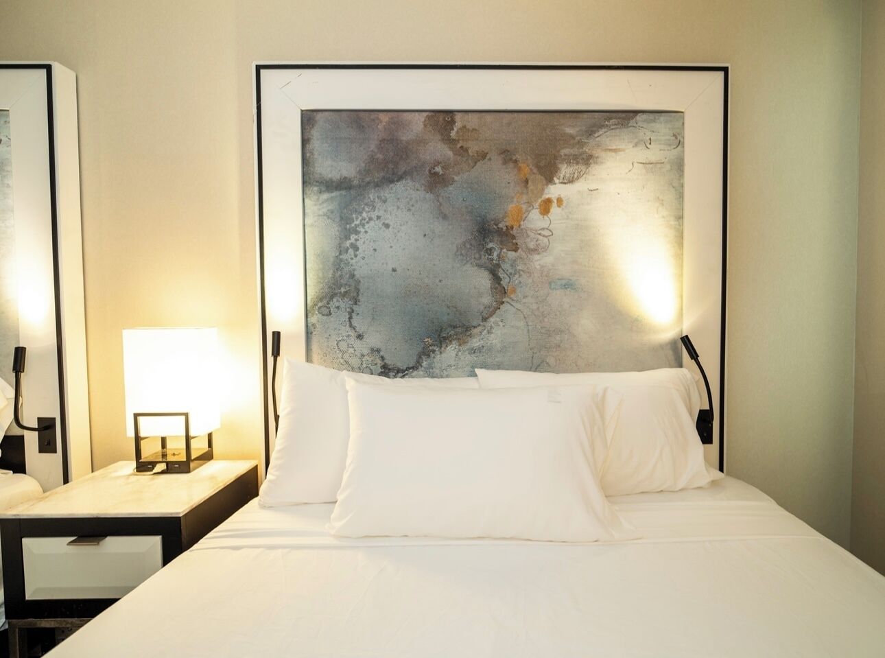 Ein Hotelzimmer mit einem großen abstrakten Gemälde an der Wand, einem ordentlich bezogenen Bett mit weißen Leinen und zwei Nachttischlampen.