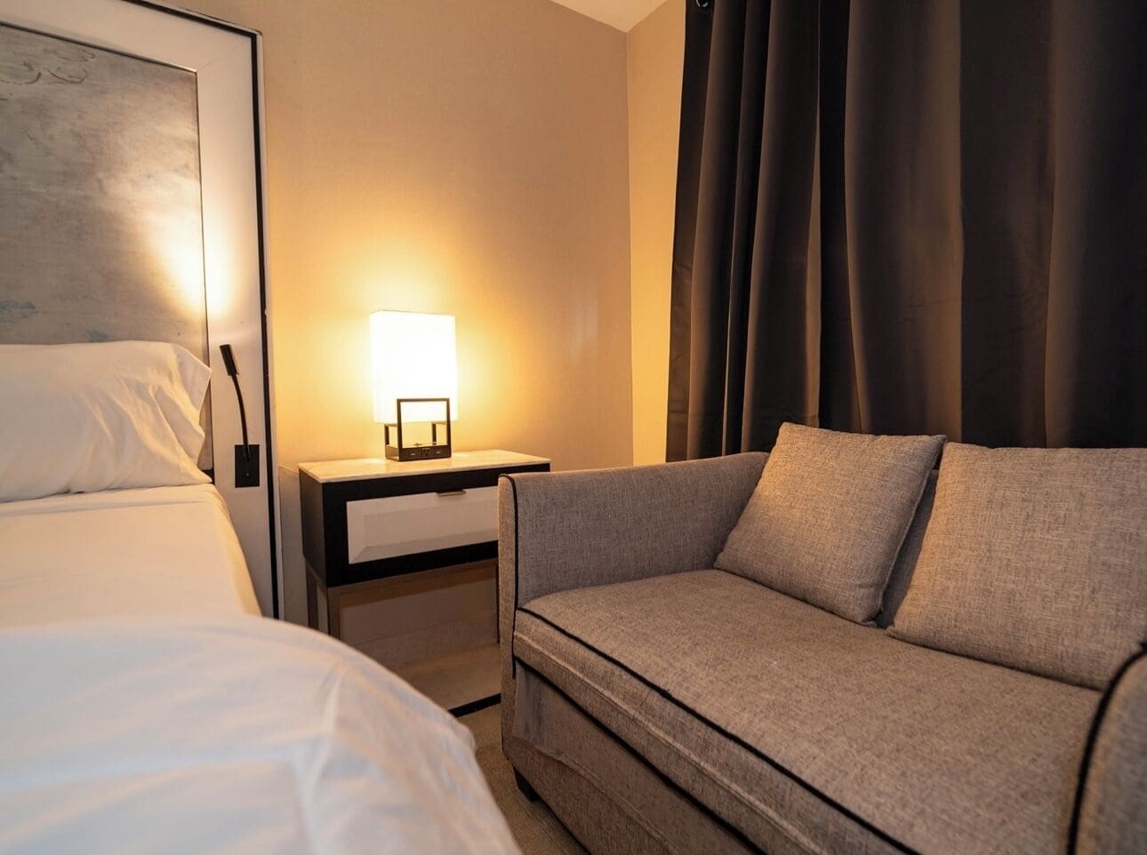 Quarto de hotel com cama, sofá, mesa de cabeceira com abajur e janela com cortinas.