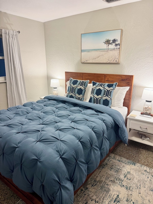 A bedroom with a large bed, wooden headboard, two bedside tables, a nightstand, a painting of a beach scene, and a patterned rug.