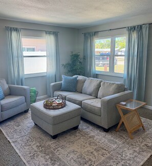 A living room with a grey sofa, a matching ottoman, a wooden side table, and a patterned rug.