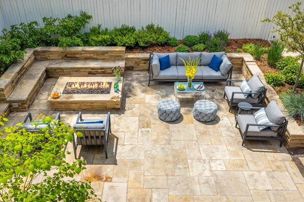 A patio with stone flooring, a fire pit, a gray sofa with blue cushions, and a coffee table with a vase of flowers.