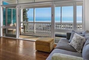 A room with a wooden floor, a grey sofa, a mustard-colored ottoman, and a view of the ocean through large sliding glass doors.
