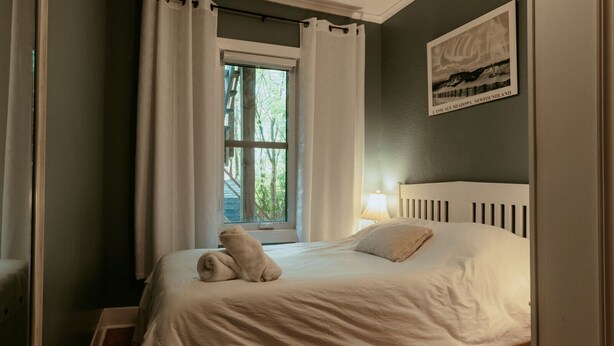 Una cama bien hecha con una almohada y una toalla enrollada, una ventana con cortinas, una lámpara en la mesita de noche y un cuadro en la pared.