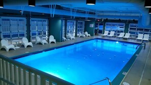 Una piscina interior con sillas reclinables blancas y vistas al mar a través de grandes ventanales.